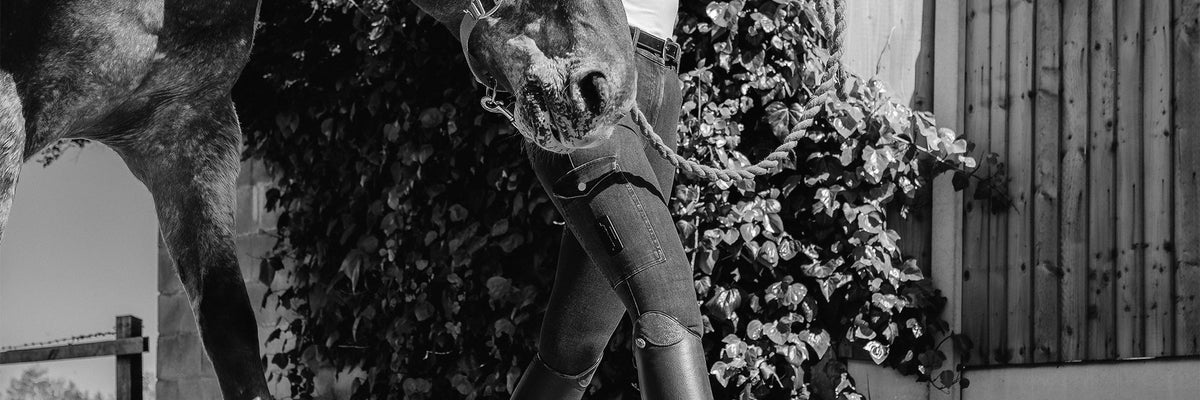 Rider in denim-style breeches and tall boots leading a horse, black and white